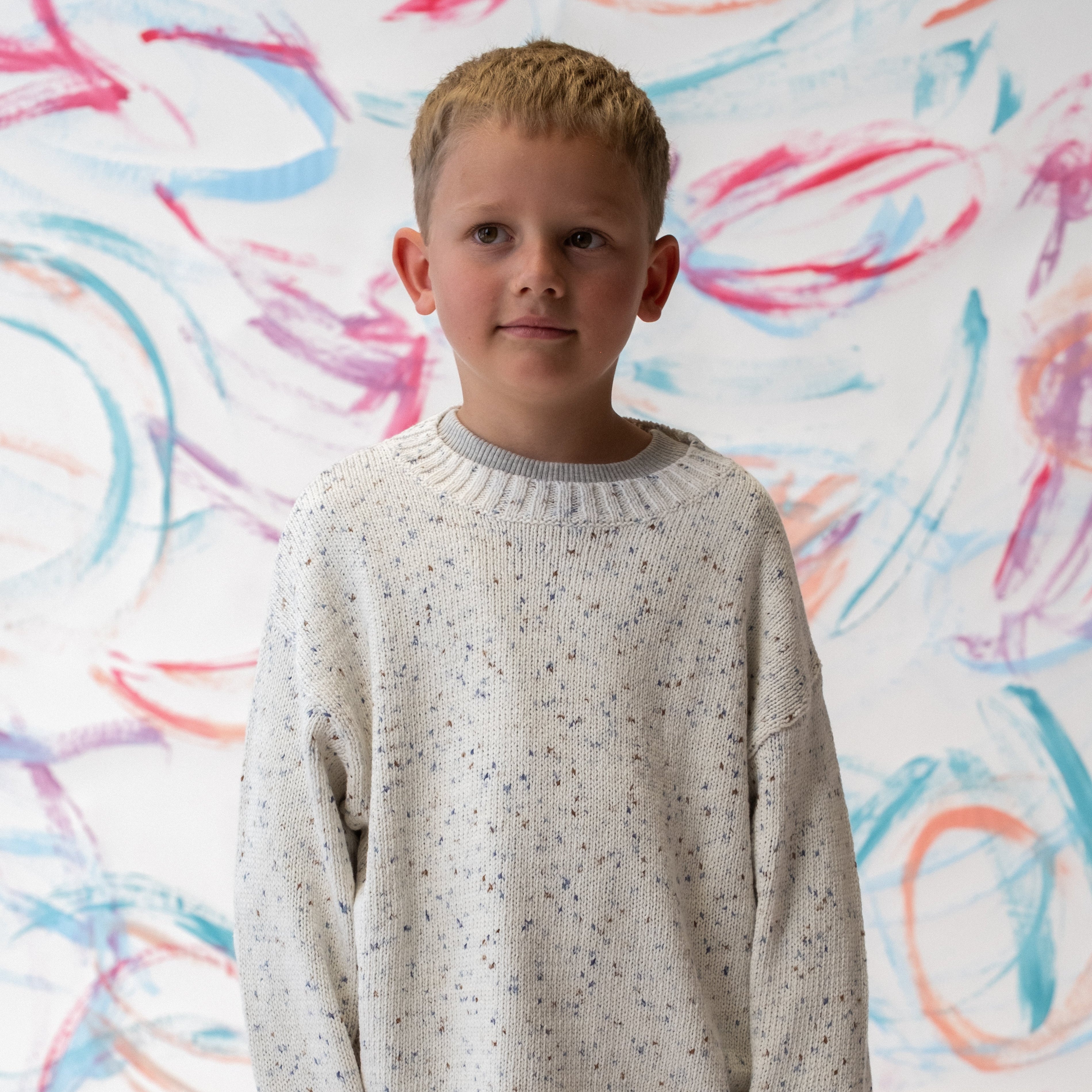 Young boy wearing a light funfetti sweater in surf against a colorful abstract background