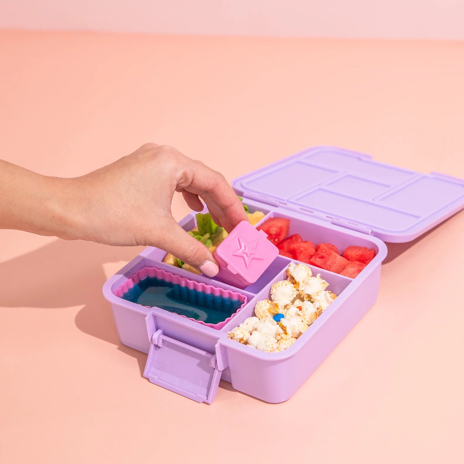 Purple bento box with compartments filled with snacks on a pink background