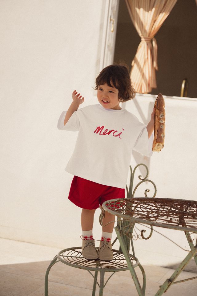Child wearing a white shirt with 'Merci' in red text, standing on a metal chair outdoors. Merci collection by White Fox & Co