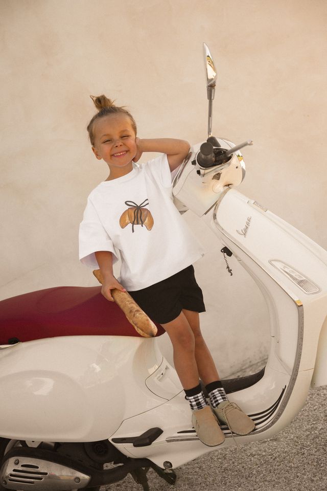 Child sitting on a scooter wearing a white t-shirt with a crossiant design by Cinnamon Baby